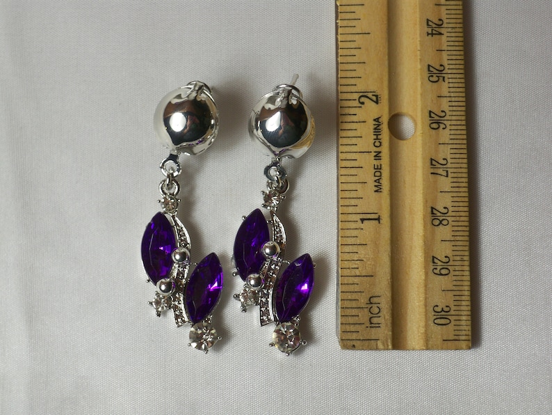 May include: A pair of silver dangle earrings with purple and clear rhinestones. The earrings are approximately 1 inch long.