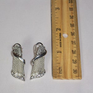 May include: A pair of silver-toned earrings with a decorative design. The earrings feature a combination of small, clear crystals and faux pearls. A wooden ruler is next to the earrings, showing inches.