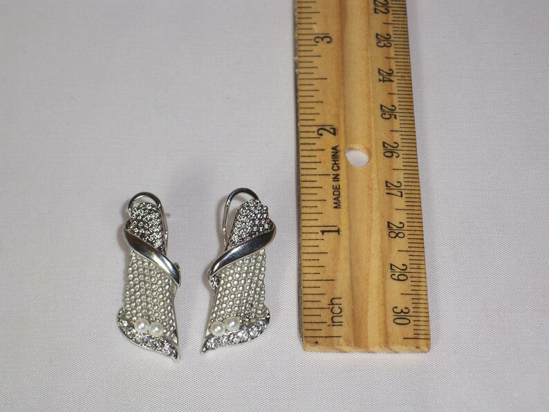 May include: A pair of silver-toned earrings with a decorative design. The earrings feature a combination of small, clear crystals and faux pearls. A wooden ruler is next to the earrings, showing inches.