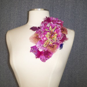 Peut inclure: Une embellissement floral rose et or avec des paillettes irisées et des plumes. L'embellissement est sur un buste de mannequin blanc.