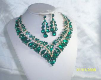May include: A green and gold necklace and earrings set with large teardrop-shaped rhinestones. The jewelry is on a white background.