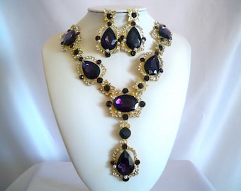 Può includere: Un set di collana e orecchini in oro con grandi pietre preziose viola a forma di goccia. La collana ha un lungo ciondolo con una grande pietra preziosa viola.