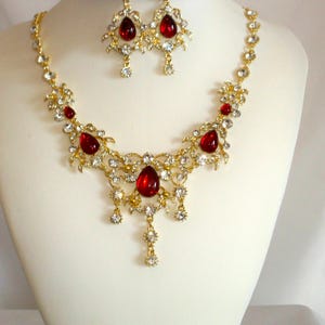 May include: A gold-toned necklace and earring set displayed on a mannequin. The necklace features large, teardrop-shaped red gemstones and smaller clear crystals. The earrings mirror the necklace design. The jewelry is ornate and likely for special occasions.
