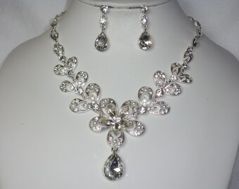 May include: A silver necklace and earrings set with a floral design and clear rhinestones. The necklace has a teardrop-shaped pendant.
