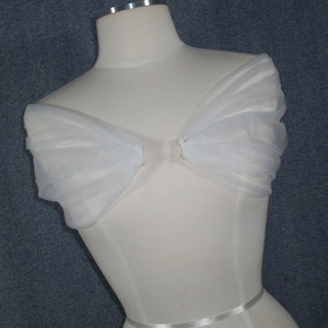 May include: White tulle fabric draped over a dress form, creating a soft and flowing look. The fabric is gathered at the shoulders and falls down the back in a cascade of ruffles.