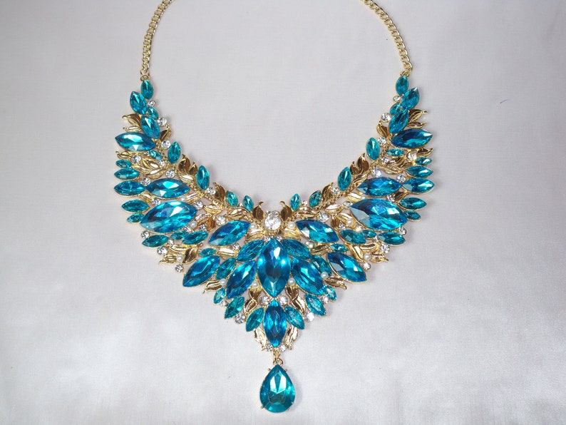 May include: A statement necklace featuring a gold-tone chain and setting. The necklace is adorned with large, vibrant blue-green gemstones and smaller clear crystals. A teardrop-shaped stone dangles from the center.