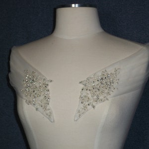 May include: A pair of white tulle shoulder straps with silver bead embellishments. The straps are designed to be worn over a wedding dress or other formal attire.