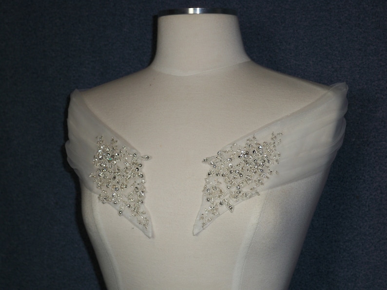 May include: A pair of white tulle shoulder straps with silver bead embellishments. The straps are designed to be worn over a wedding dress or other formal attire.