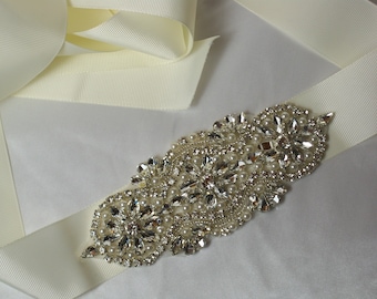 May include: A white satin ribbon with a large, ornate rhinestone and pearl embellishment. The embellishment is silver and features a cluster of rhinestones and pearls in a floral design.