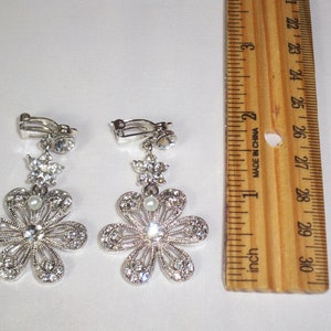 May include: A pair of silver-toned clip-on earrings featuring a flower design with clear rhinestones and a small white pearl at the centre. A wooden ruler is placed next to the earrings, showing they measure approximately 6.4 cm in length.