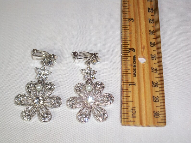 May include: A pair of silver-toned clip-on earrings featuring a flower design with clear rhinestones and a small white pearl at the centre. A wooden ruler is placed next to the earrings, showing they measure approximately 6.4 cm in length.