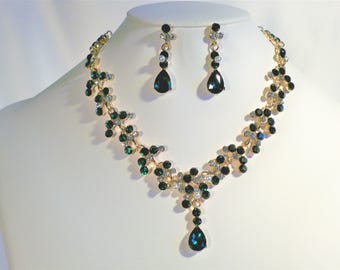 May include: A gold-toned necklace and earring set displayed on a mannequin. The necklace and earrings feature dark green and clear gemstones, with a teardrop-shaped pendant on the necklace. The jewelry is designed with a floral vine pattern.