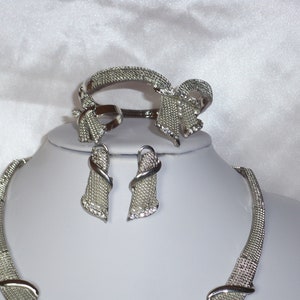 May include: A silver-toned jewelry set featuring a necklace, bracelet, ring, and earrings. The set includes a mesh-like design with bow accents and small, sparkling embellishments. The jewelry is displayed on a white mannequin.