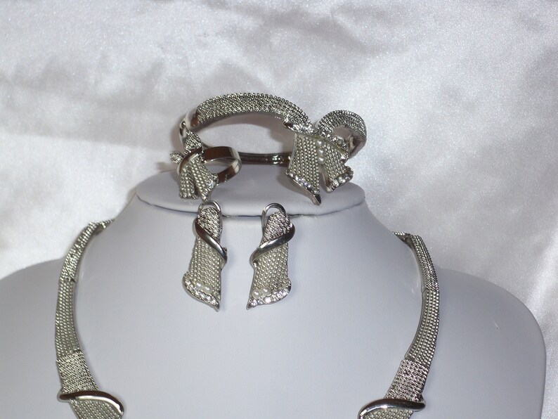 May include: A silver-toned jewelry set featuring a necklace, bracelet, ring, and earrings. The set includes a mesh-like design with bow accents and small, sparkling embellishments. The jewelry is displayed on a white mannequin.