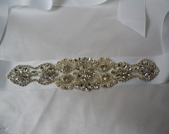 May include: A white satin ribbon with a decorative embellishment of rhinestones and pearls. The embellishment is in the shape of a floral design.