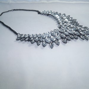 Puede incluir: Un collar llamativo con una cadena de metal oscuro y un dise&ntilde;o de cuello ancho y superpuesto, adornado con numerosos cristales transparentes de talla marquesa dispuestos en un patr&oacute;n denso similar a hojas.