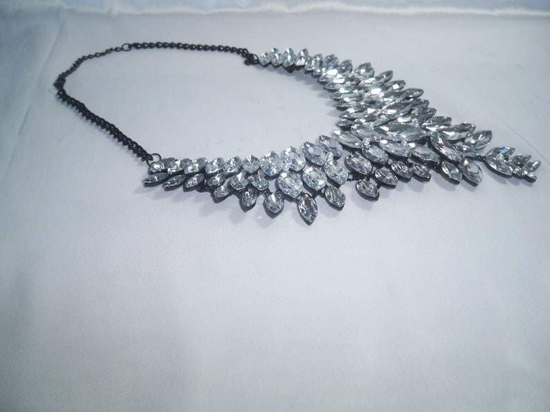 Puede incluir: Un collar llamativo con una cadena de metal oscuro y un dise&ntilde;o de cuello ancho y superpuesto, adornado con numerosos cristales transparentes de talla marquesa dispuestos en un patr&oacute;n denso similar a hojas.