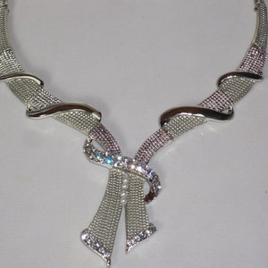 May include: Silver-toned necklace with a bow-shaped pendant. The necklace features a mesh design with sparkling rhinestones and a few small pearls. The necklace has a curved design and is suitable for formal occasions.