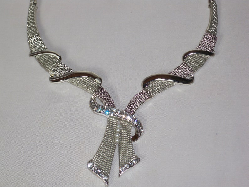 May include: Silver-toned necklace with a bow-shaped pendant. The necklace features a mesh design with sparkling rhinestones and a few small pearls. The necklace has a curved design and is suitable for formal occasions.