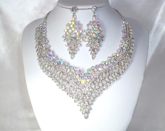 Op de afbeelding: Een zilverkleurige ketting- en oorbellenset. De ketting heeft een trapsgewijs ontwerp met iriserende kristallen. De bijpassende oorbellen zijn een cluster van vergelijkbare kristallen. De sieraden worden getoond op een witte mannequin.