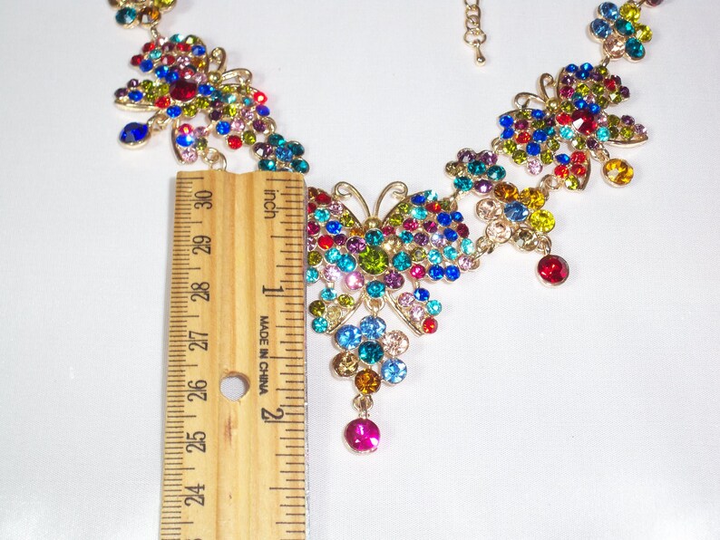 Puede incluir: Un collar de mariposa colorido con una cadena dorada y una variedad de piedras de cristal de colores. El collar mide aproximadamente 5 cm de largo.