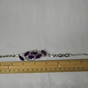 May include: A silver bracelet with a purple rhinestone design. The bracelet is 7 inches long and has a chain link style.