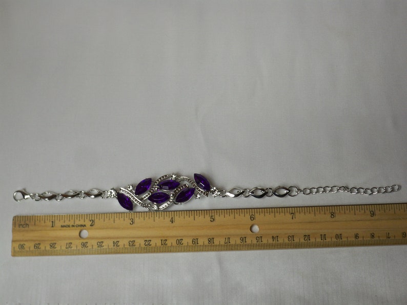 May include: A silver bracelet with a purple rhinestone design. The bracelet is 7 inches long and has a chain link style.