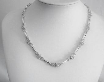 May include: A silver-toned necklace featuring a repeating pattern of smooth, elongated metallic bars alternating with small, circular clusters of clear, sparkling crystals. The design creates a continuous, symmetrical chain around the neck.
