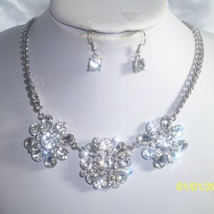 May include: A silver necklace with three flower-shaped pendants made of clear rhinestones. The necklace is paired with matching earrings with a single clear rhinestone.