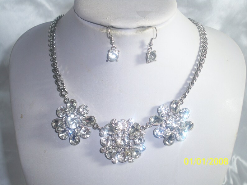 May include: A silver necklace with three flower-shaped pendants made of clear rhinestones. The necklace is paired with matching earrings with a single clear rhinestone.