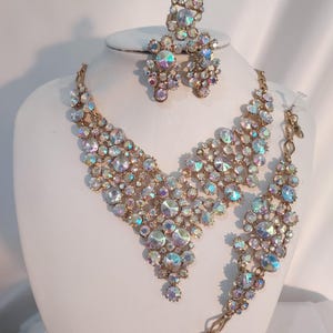 May include: A gold-toned jewelry set featuring a necklace, bracelet, earrings, and ring. The set is adorned with iridescent, clear, and colorful rhinestones. The jewelry is displayed on a white mannequin head.