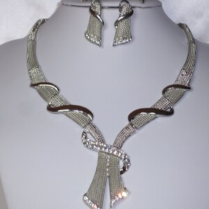 May include: Silver-toned necklace and earring set. The necklace features a mesh design with curved accents and a central bow-like embellishment adorned with clear crystals. The matching earrings have a similar mesh design.