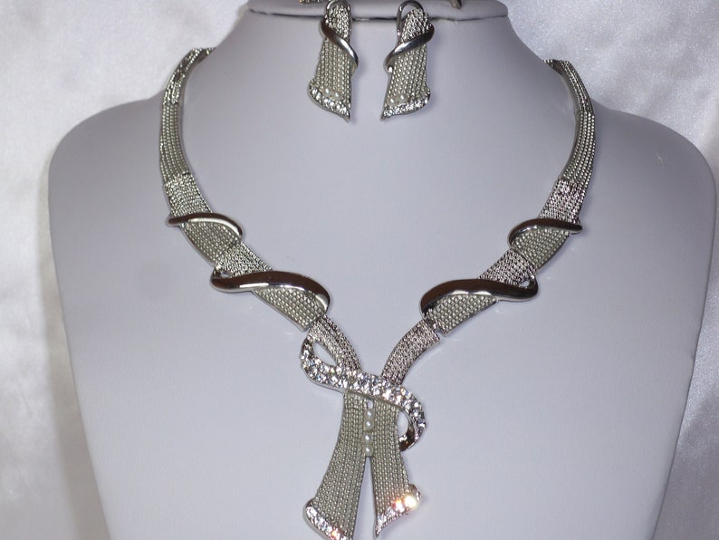 May include: Silver-toned necklace and earring set. The necklace features a mesh design with curved accents and a central bow-like embellishment adorned with clear crystals. The matching earrings have a similar mesh design.