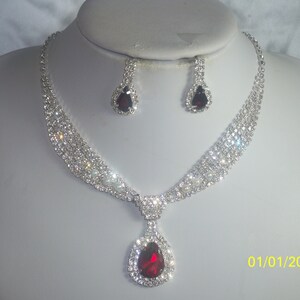 May include: A silver necklace with a teardrop-shaped pendant set with red and clear rhinestones. The necklace is paired with matching earrings.