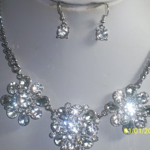 May include: A silver necklace with three flower-shaped pendants made of clear rhinestones. The necklace is paired with matching earrings with a single clear rhinestone.