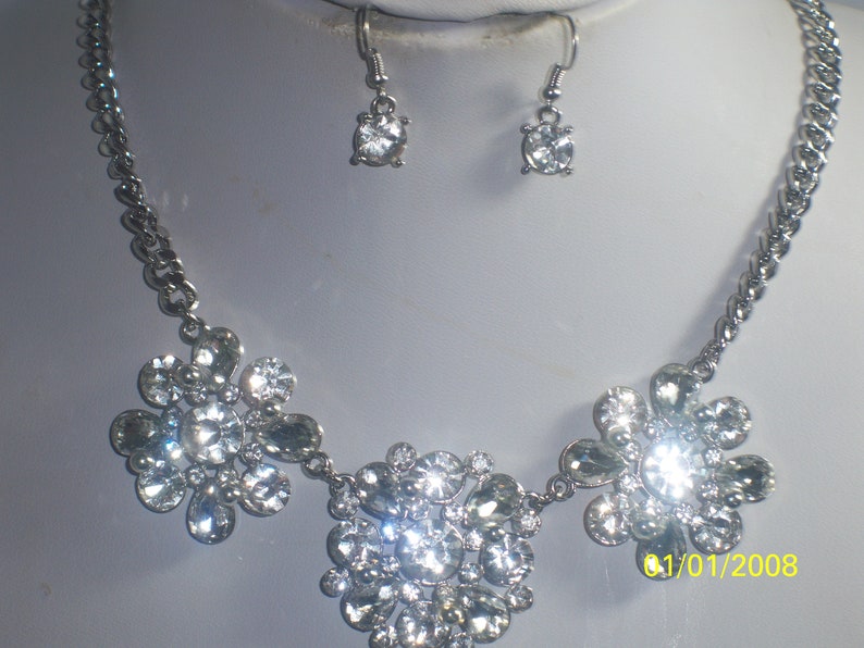 May include: A silver necklace with three flower-shaped pendants made of clear rhinestones. The necklace is paired with matching earrings with a single clear rhinestone.