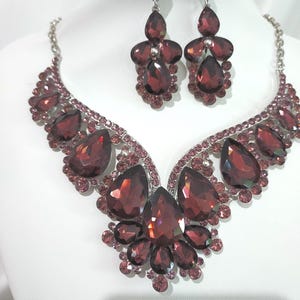 May include: A burgundy-colored jewelry set featuring a necklace and matching earrings. The necklace has a silver-toned chain and is adorned with large, teardrop-shaped and round burgundy crystals. The earrings mirror the necklace design with similar crystals.