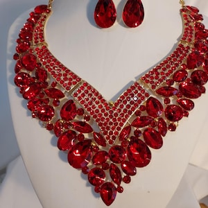 May include: A gold-tone necklace and matching drop earring set featuring numerous faceted red crystals in various shapes and sizes. The necklace has a V-shaped design with a dense arrangement of round and teardrop-shaped red stones set in metal.