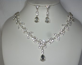May include: A silver necklace and earrings set with a teardrop-shaped crystal pendant and smaller crystals in a floral design.
