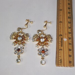 May include: A pair of gold tone clip-on earrings with a butterfly design. The butterflies are adorned with clear and amber rhinestones. Each earring has a dangling flower charm with a clear and amber rhinestone design. The earrings are approximately 2 inches long.