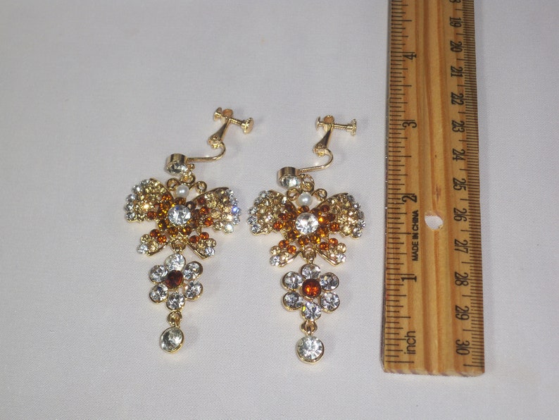 May include: A pair of gold tone clip-on earrings with a butterfly design. The butterflies are adorned with clear and amber rhinestones. Each earring has a dangling flower charm with a clear and amber rhinestone design. The earrings are approximately 2 inches long.