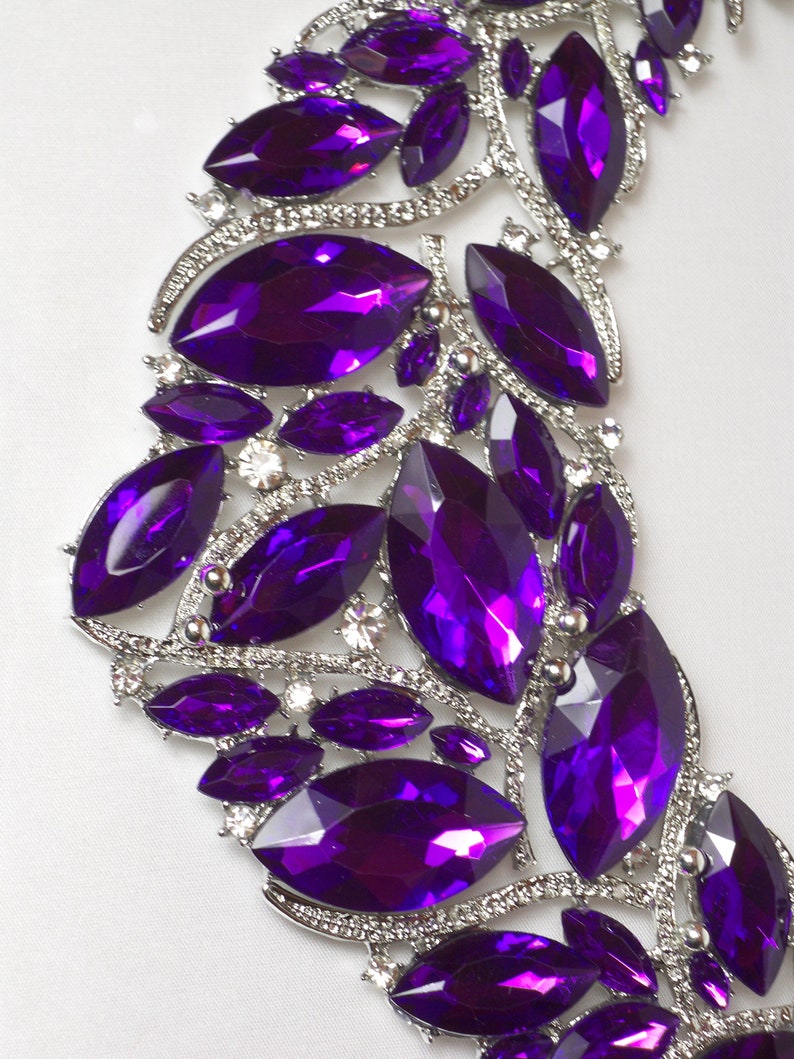 May include: A silver and purple jeweled necklace with a cascading design. The necklace is made of metal and features a variety of purple rhinestones in different sizes and shapes.