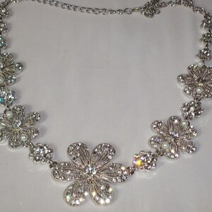 May include: A silver-toned necklace featuring a series of connected flower-shaped links. Each flower is encrusted with clear, sparkling rhinestones and accented with a small, round white pearl at the centre. The necklace includes a delicate chain.