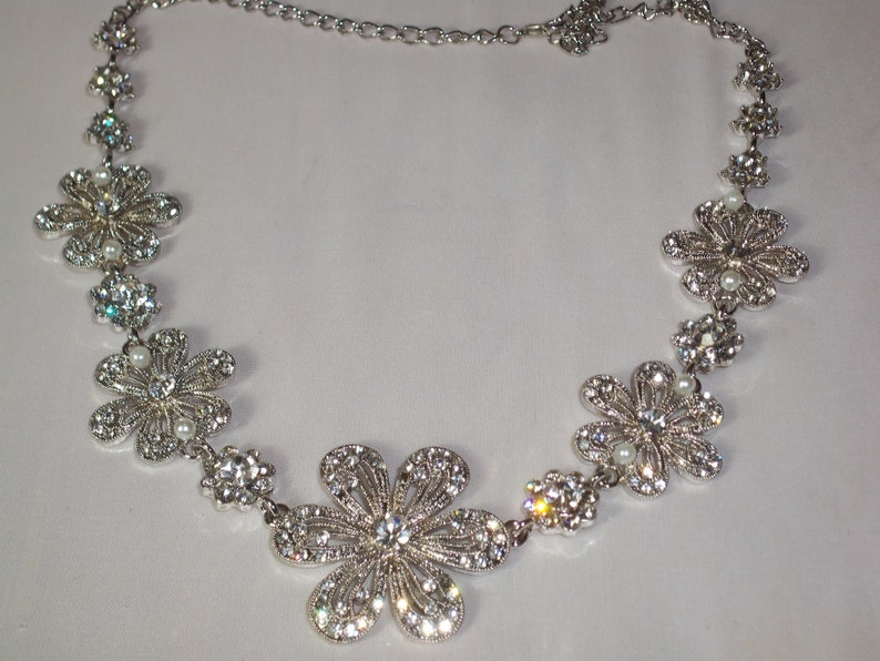 May include: A silver-toned necklace featuring a series of connected flower-shaped links. Each flower is encrusted with clear, sparkling rhinestones and accented with a small, round white pearl at the centre. The necklace includes a delicate chain.