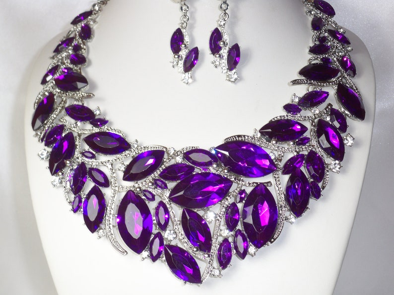 May include: A silver necklace and earrings set with large, purple, marquise-shaped rhinestones. The necklace is a statement piece with a cascading design.