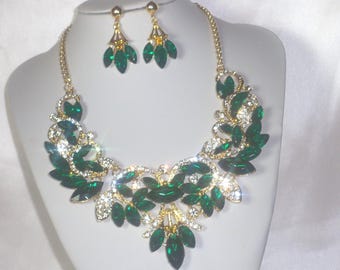 May include: A gold-tone necklace and matching drop earring set featuring marquise-cut dark green crystals and clear round rhinestones arranged in a leaf-like pattern. The necklace has a thick chain and a central cluster of green and clear stones.