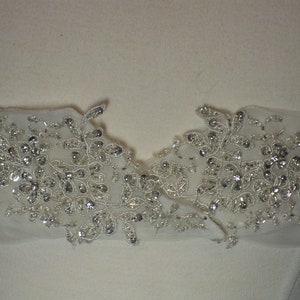 May include: Close-up of a white fabric with silver beaded floral embellishments. The embellishments are arranged in a symmetrical pattern, creating a delicate and elegant design.