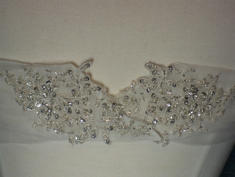 May include: Close-up of a white fabric with silver beaded floral embellishments. The embellishments are arranged in a symmetrical pattern, creating a delicate and elegant design.