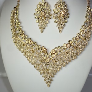 May include: A gold-tone necklace and earrings set with a delicate floral design and clear rhinestones. The necklace has a V-shaped pendant and the earrings are teardrop shaped.