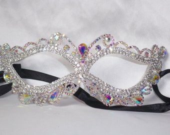 May include: A silver masquerade mask with iridescent rhinestones and pearl accents. The mask has a black ribbon attached for wearing.